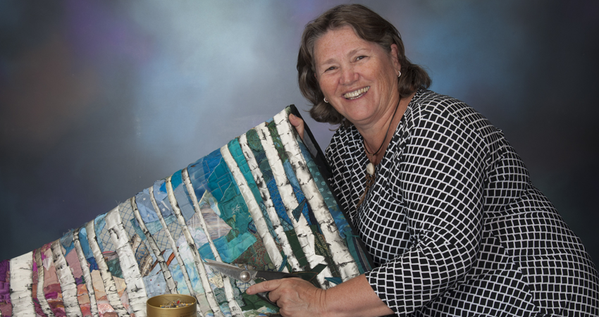 Smiling woman displays a colorful quilt against a gradient backdrop.